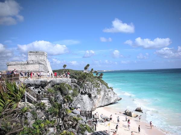 Ancient Mayan stone temple on Tulum cliff, tourists on Caribbean beach below