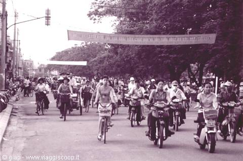 Cyclists and mopeds ride down a tree-lined avenue under banners