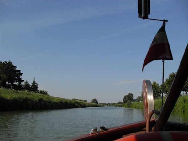 Wooden boat with red and white flag cruising a canal between grassy banks