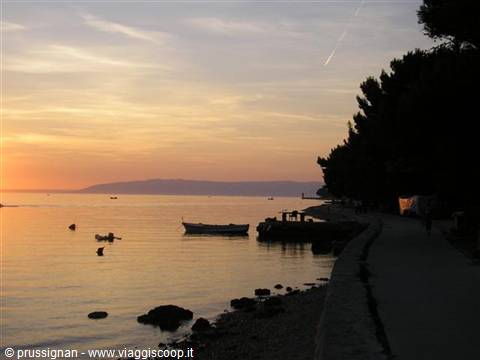 Boats on calm sea, tree-lined promenade under golden evening sky