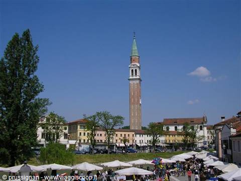 Leaning brick bell tower with green spire above rooftops, market umbrellas below