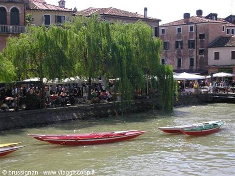 Red boats moored on a Venice canal, crowded outdoor cafés under willows