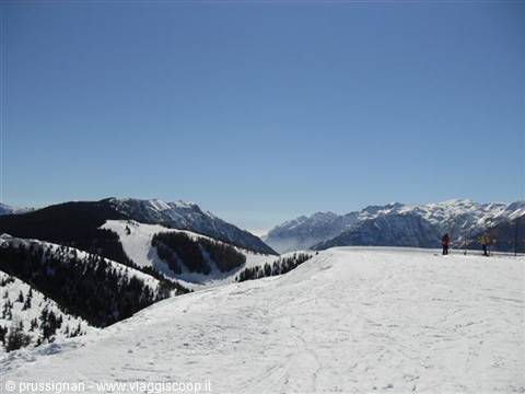 Snowy ski slope between forested mountains, skier in distance, clear blue sky