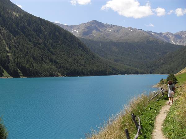 Woman walks on dirt path beside turquoise alpine lake among forested slopes