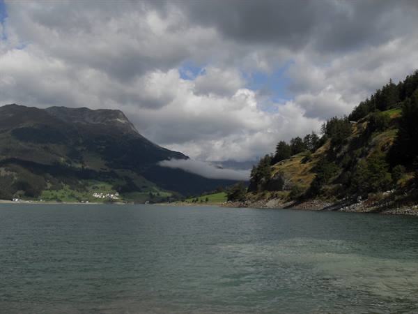 Alpine lake with white village on shore, green mountains and low clouds