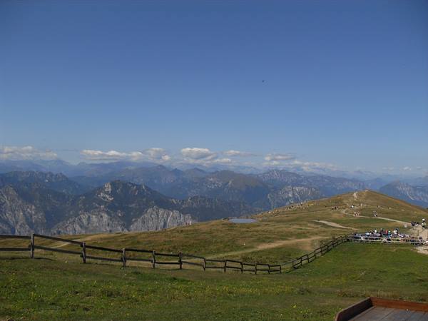 Wooden fence on alpine meadow, tourists walking toward mountains under blue sky