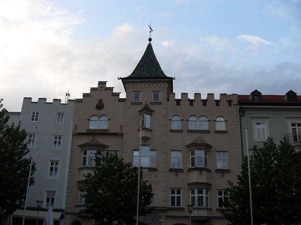 Crenellated building with conical tower under cloudy sky, trees in front