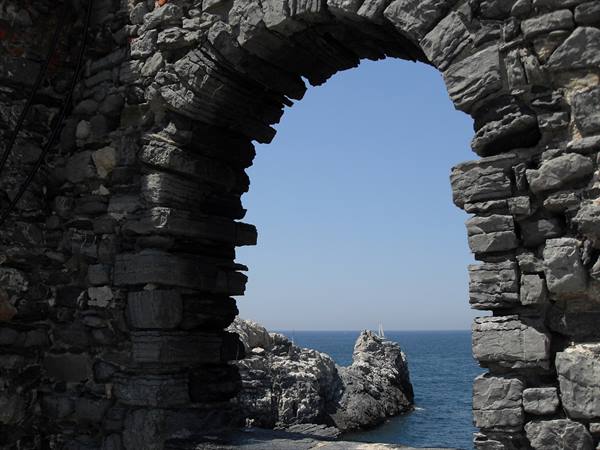 Grey stone arch framing rocks and blue sea, a sailboat in the distance