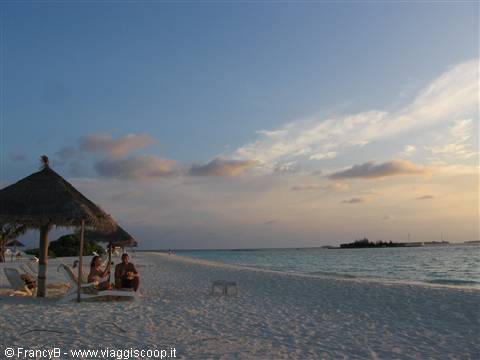 Couple on loungers under thatched umbrella, white sand beach at dusk