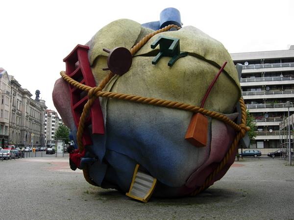 Giant sphere sculpture tied with rope, covered with ladders and objects, in city square