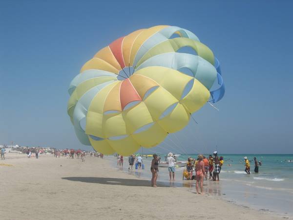 Beachgoers on the sand prepare a huge yellow parasailing canopy under blue sky