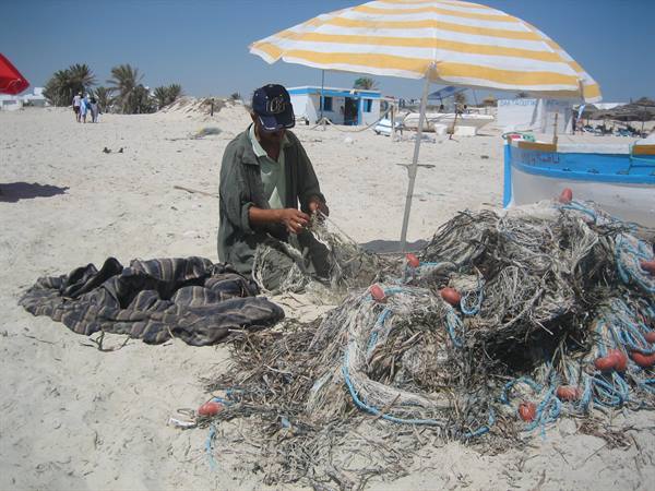 Fisherman mends nets on sandy beach, under yellow and blue umbrella, beside his boat