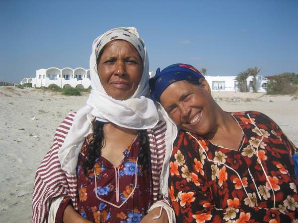 Two smiling women wearing headscarves on sandy beach, white houses behind