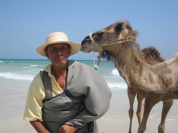 Man in straw hat standing beside a camel on a sunny beach