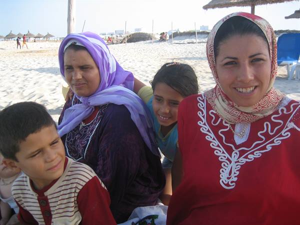 Women in headscarves with children on white sand beach, straw umbrellas behind