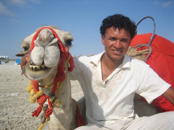 Smiling young man beside a camel with red reins on the sand