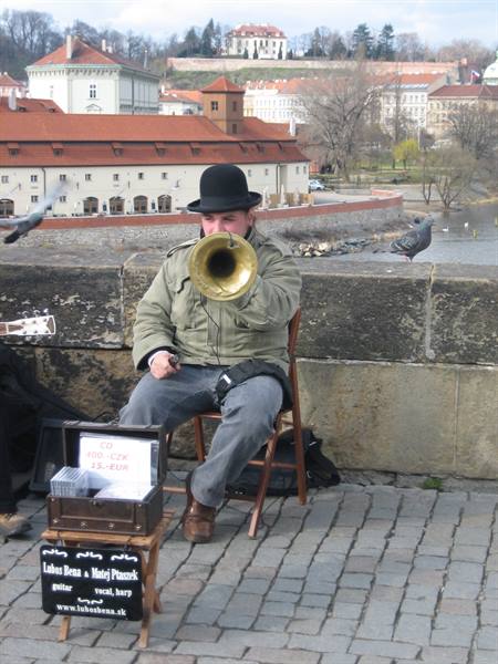 Musician in bowler hat plays flugelhorn on Charles Bridge, pigeons and Prague rooftops
