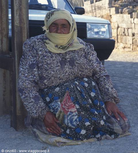 Woman in yellow headscarf sitting on the ground beside a car, stone walls
