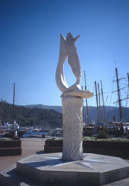 Marble mermaid and dolphin sculpture on the waterfront, sailboat masts behind