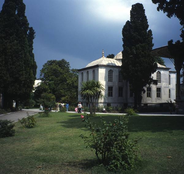 Dome-topped pavilion among cypresses, green lawn and roses, visitors strolling in Topkapi