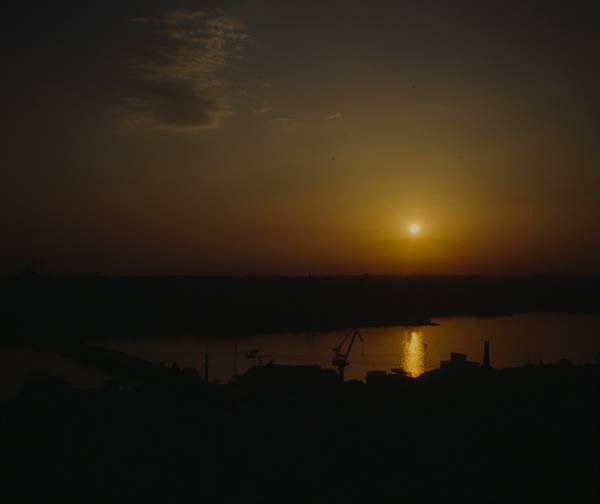 Low sun glowing over a dark river with cranes and silhouetted buildings
