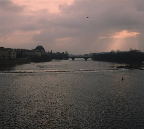River with a weir, a stone bridge and birds under cloudy dusk sky in Prague