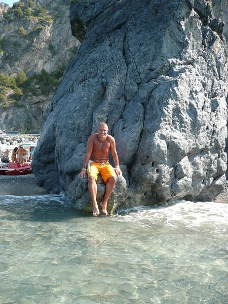 Man in orange swimwear sitting on large grey rock, clear water and beach