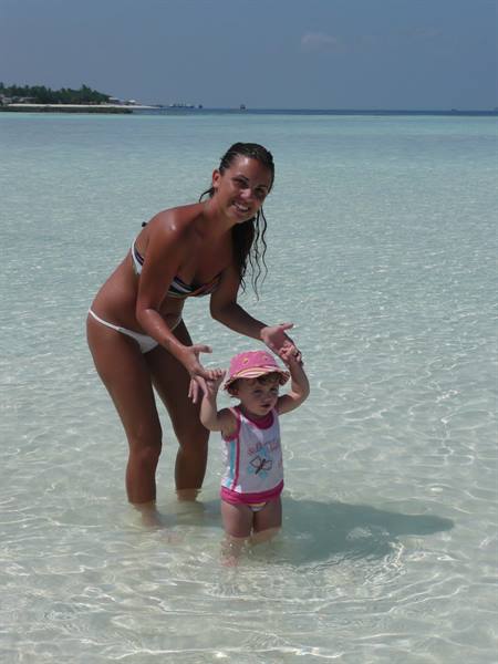 Mother in bikini helps toddler take steps in shallow turquoise water