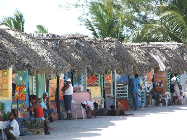 Thatched-roof craft stalls on tropical beach, painted artwork vendors and tourists in sunlight
