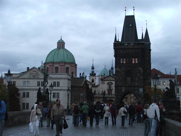 Tourists walk on Charles Bridge toward the Gothic tower, under Prague's grey sky