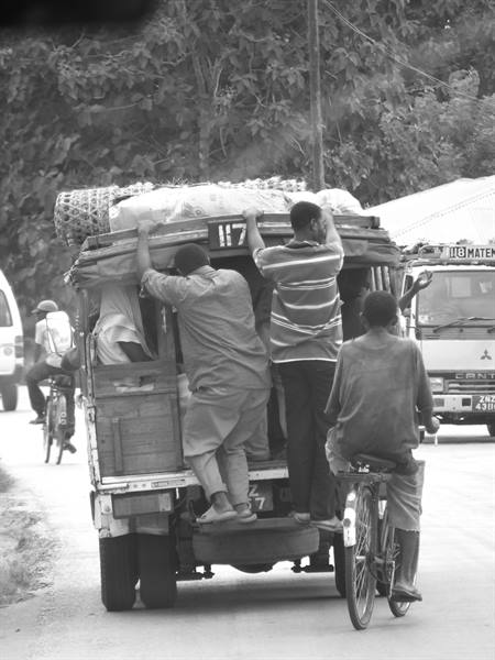 Men clinging to a loaded minibus, cyclist beside, dirt road traffic