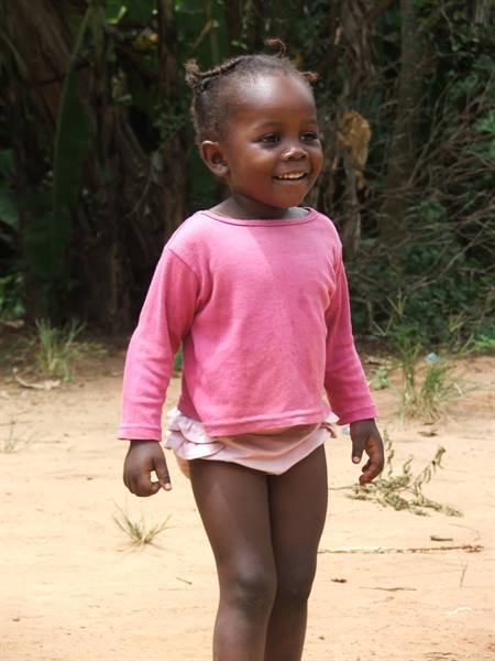 Smiling little girl in pink shirt standing on sandy ground with green trees