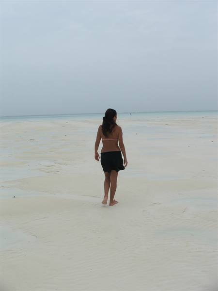 Barefoot woman walks on white sand toward the sea under grey sky