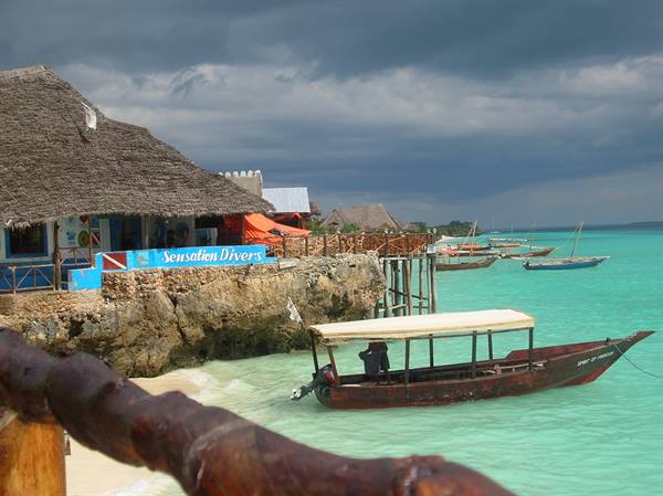Thatched-roof dive bar on rocks, wooden boat moored in turquoise water, Nungwi