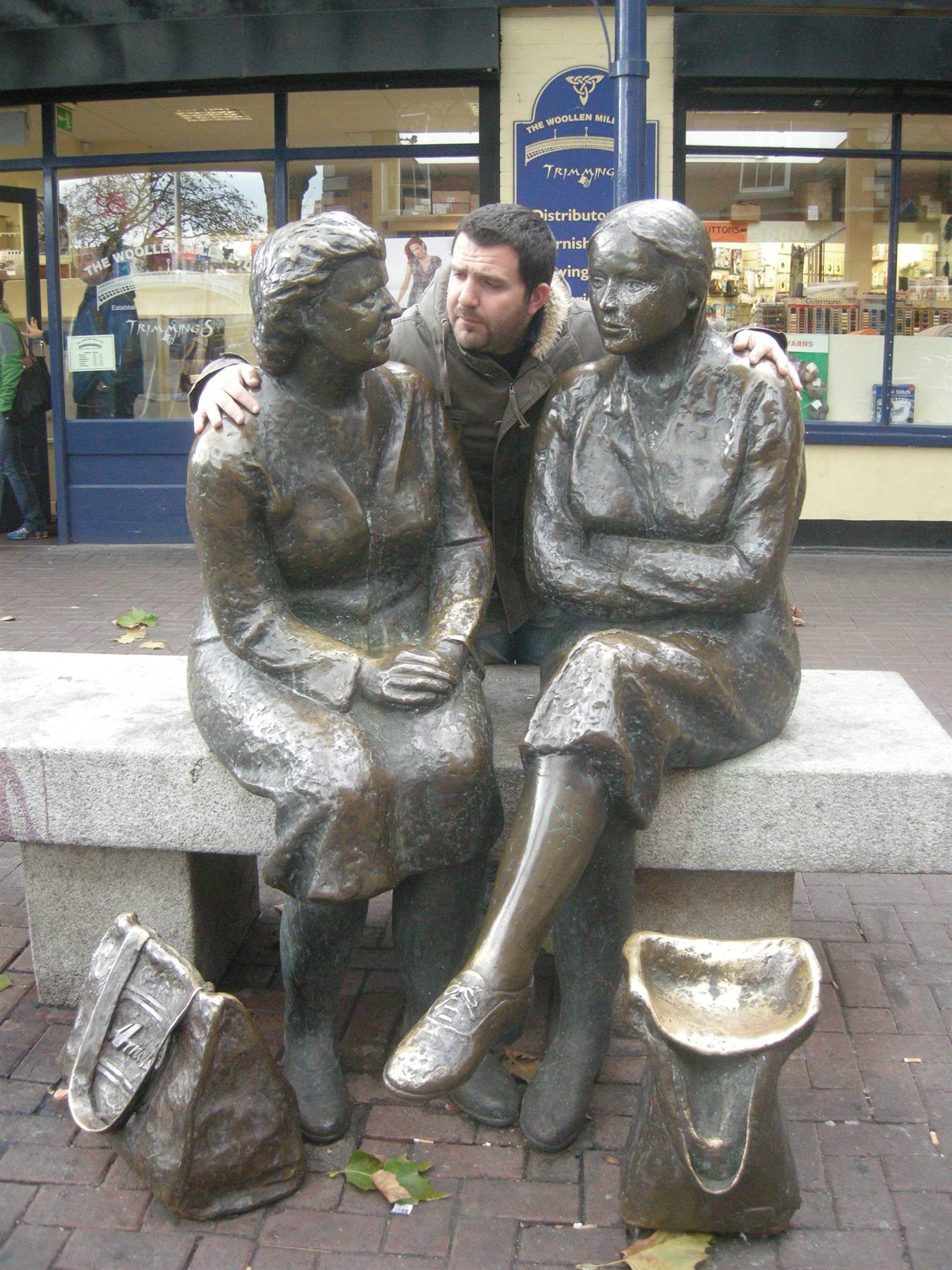 A man embraces two bronze statues of seated women from behind on a bench