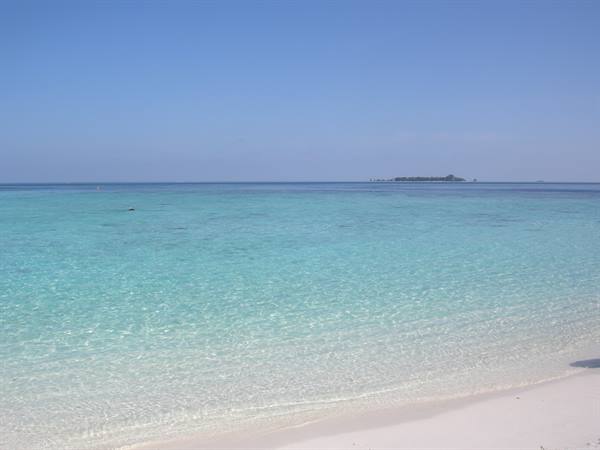 Clear turquoise water on white sand, palm island on the horizon
