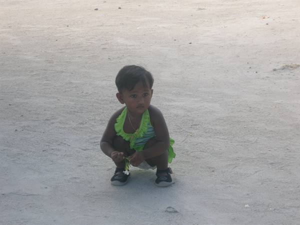 Toddler squatting on white sand, green dress, holding a small flower