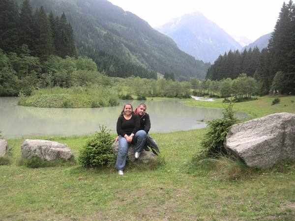 Two people sitting on a boulder by a small lake, fir trees and mountains around