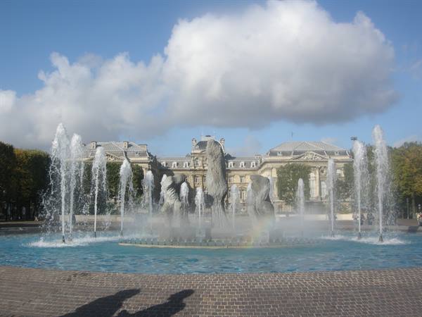 Fountain with water jets and sculptures before a classical building, cloudy sky
