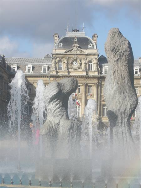 Fountain with stone sculptures and water jets before a clock-topped building