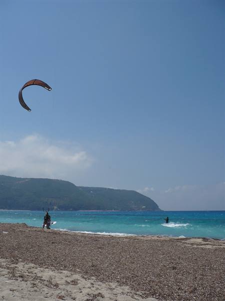 Kitesurfer on turquoise water, pebble beach, green hills in the background