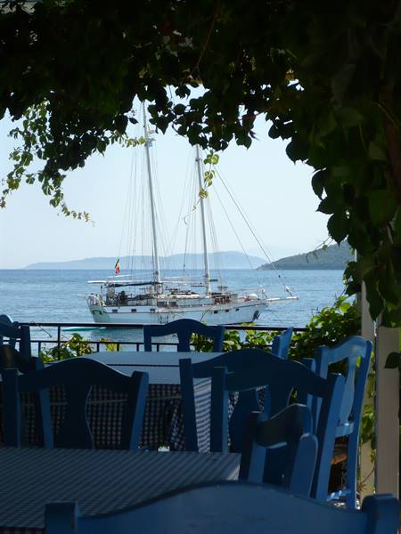 Taverna blue chairs under green pergola, white motor sailor anchored ahead