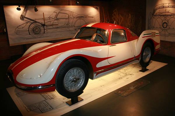 Red and white vintage racing car displayed on illuminated technical drawings