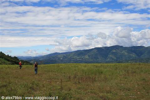 Two hikers walking across a green meadow, mountains and white clouds behind