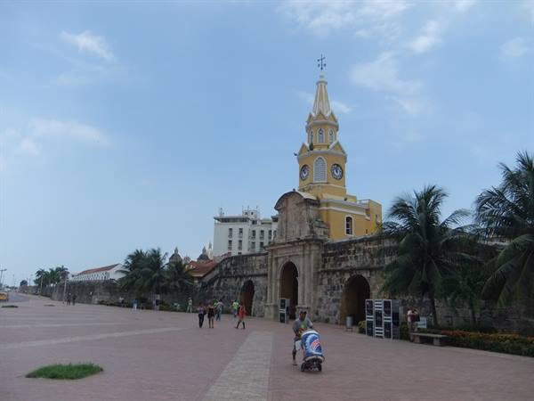 Yellow clock tower atop stone walls, plaza with street vendor, palms, Cartagena