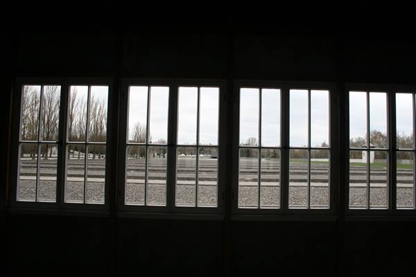 Four-paned windows of a dark barrack frame gravel grounds, bare trees and grey sky
