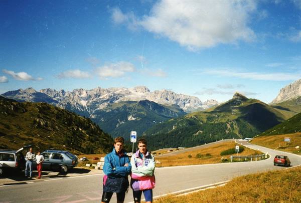Two cyclists pose on a winding Dolomites pass road, parked cars nearby