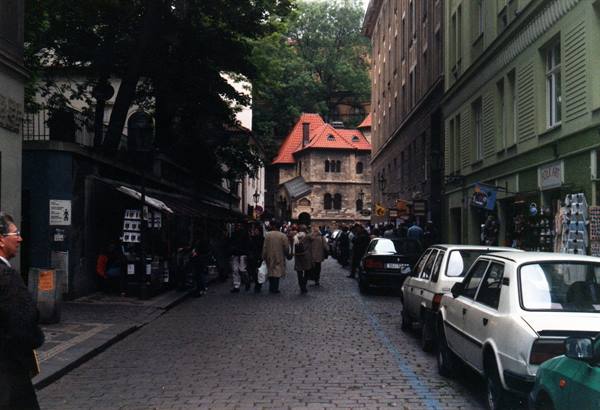 Cobbled street with pedestrians and parked cars, green houses, stone building, Prague