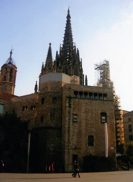 Gothic spire of Barcelona Cathedral above medieval stone walls, tourists walk below