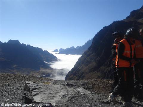 Motorcyclists in orange jackets stop on gravel road above clouds between mountains